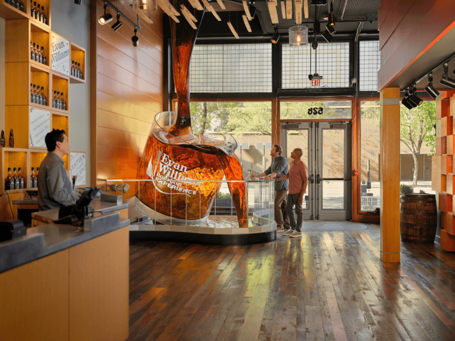 All-Ages Adventure: The Ultimate Trip to Louisville, KY - 14 Two people stand by a large display of a whiskey glass and pouring bottle inside a modern Louisville tasting room, while a staff member stands behind the counter—an inviting stop on your trip.