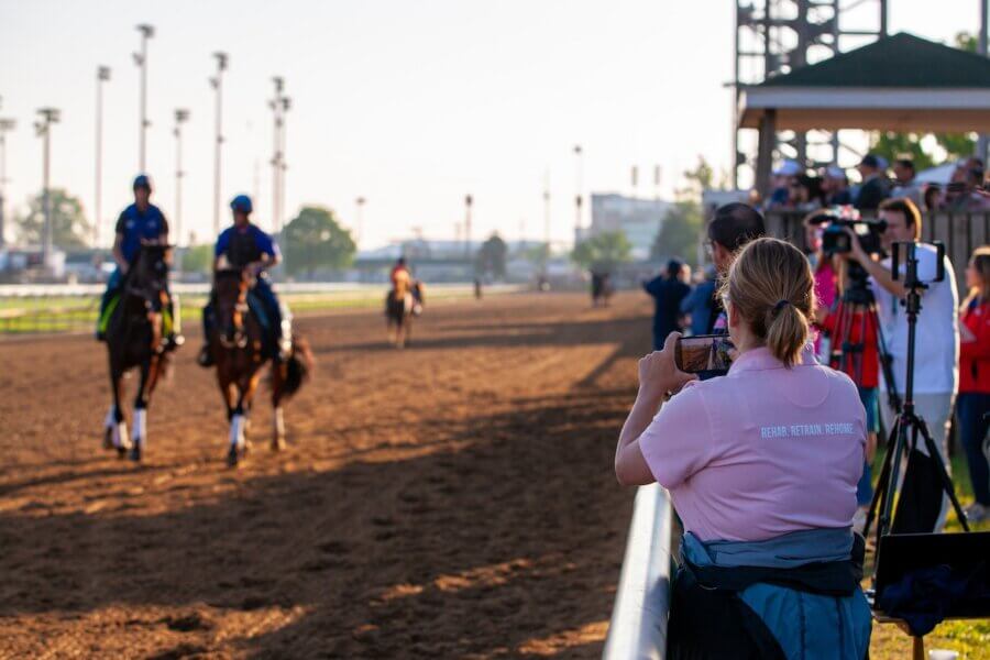 All-Ages Adventure: The Ultimate Trip to Louisville, KY - 4 A woman in a pink shirt snaps a photo with her phone of two people riding horses on a racetrack—just one of the exciting things to do in Louisville, as others stand nearby with cameras capturing the moment.