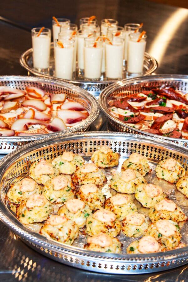 All-Ages Adventure: The Ultimate Trip to Louisville, KY - 10 Four trays of assorted appetizers on a metal surface, including stuffed pastries and sliced meats with cheese—perfect for sampling while exploring things to do in Louisville. Glasses of a light creamy beverage with pretzel sticks complete the spread.