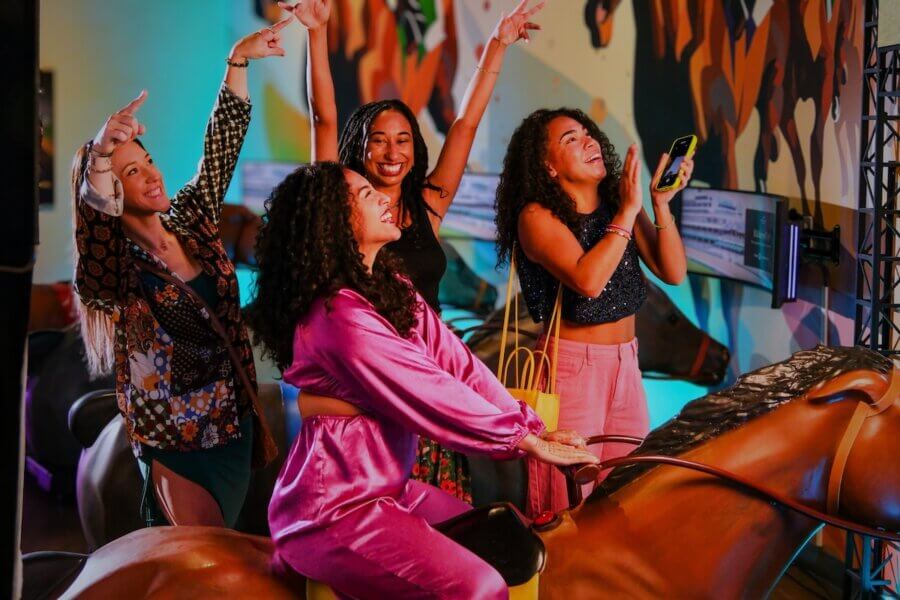 All-Ages Adventure: The Ultimate Trip to Louisville, KY - 2 Four women smile and gesture enthusiastically while posing on and around a mechanical horse indoors, with colorful decor and lighting in the background—a lively snapshot of fun things to do in Louisville.