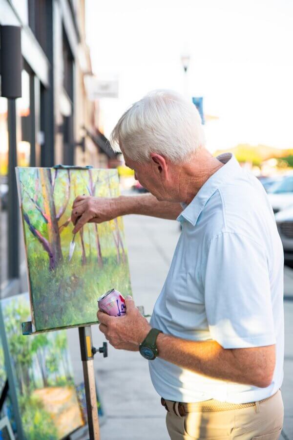 16 Can't-Miss Events in Meridian, MS This Season - 6 An older man in a light blue shirt paints a landscape on an easel outdoors during meridian events, holding a can of LaCroix sparkling water.
