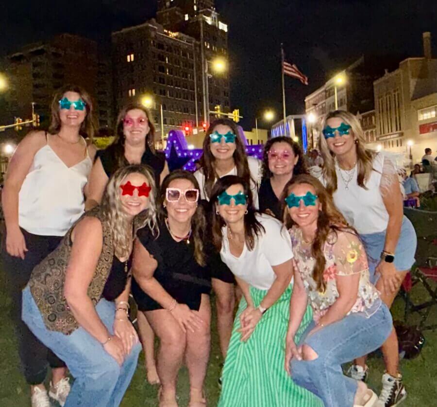 16 Can't-Miss Events in Meridian, MS This Season - 4 A group of nine women pose together outdoors at night, all wearing colorful star-shaped glasses during a lively Meridian Events gathering, with city buildings and lights twinkling in the background.