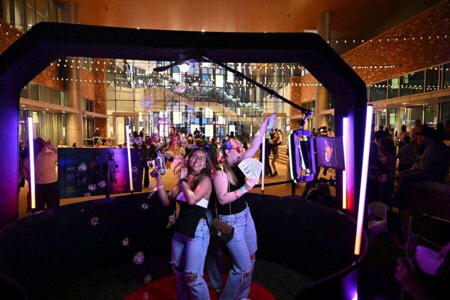 16 Can't-Miss Events in Meridian, MS This Season - 2 Two people pose and smile inside a photo booth setup by Meridian Events, surrounded by bubbles and bright lights, at a lively indoor gathering with a crowd in the background.