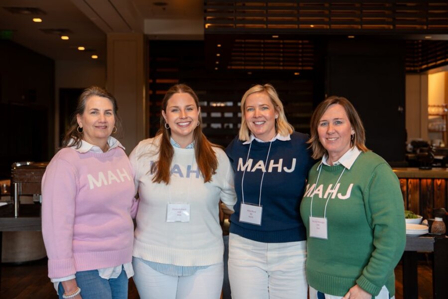 8 Mahjong Retreats Worth the Trip in 2026 - 5 Four women wearing "MAHJ" sweaters stand together in a restaurant, smiling at the cameraβcapturing the spirit of relaxation and friendship often found at Mahjong retreats in the South.
