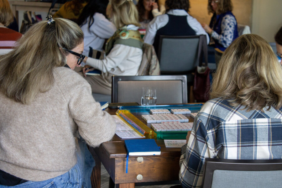 8 Mahjong Retreats Worth the Trip in 2026 - 4 Two women sit at a table playing a tile-based game, enjoying relaxation and friendly competition. Scorecards and a notebook rest nearby, evoking the spirit of Mahjong retreats in the South as others chat in the background.