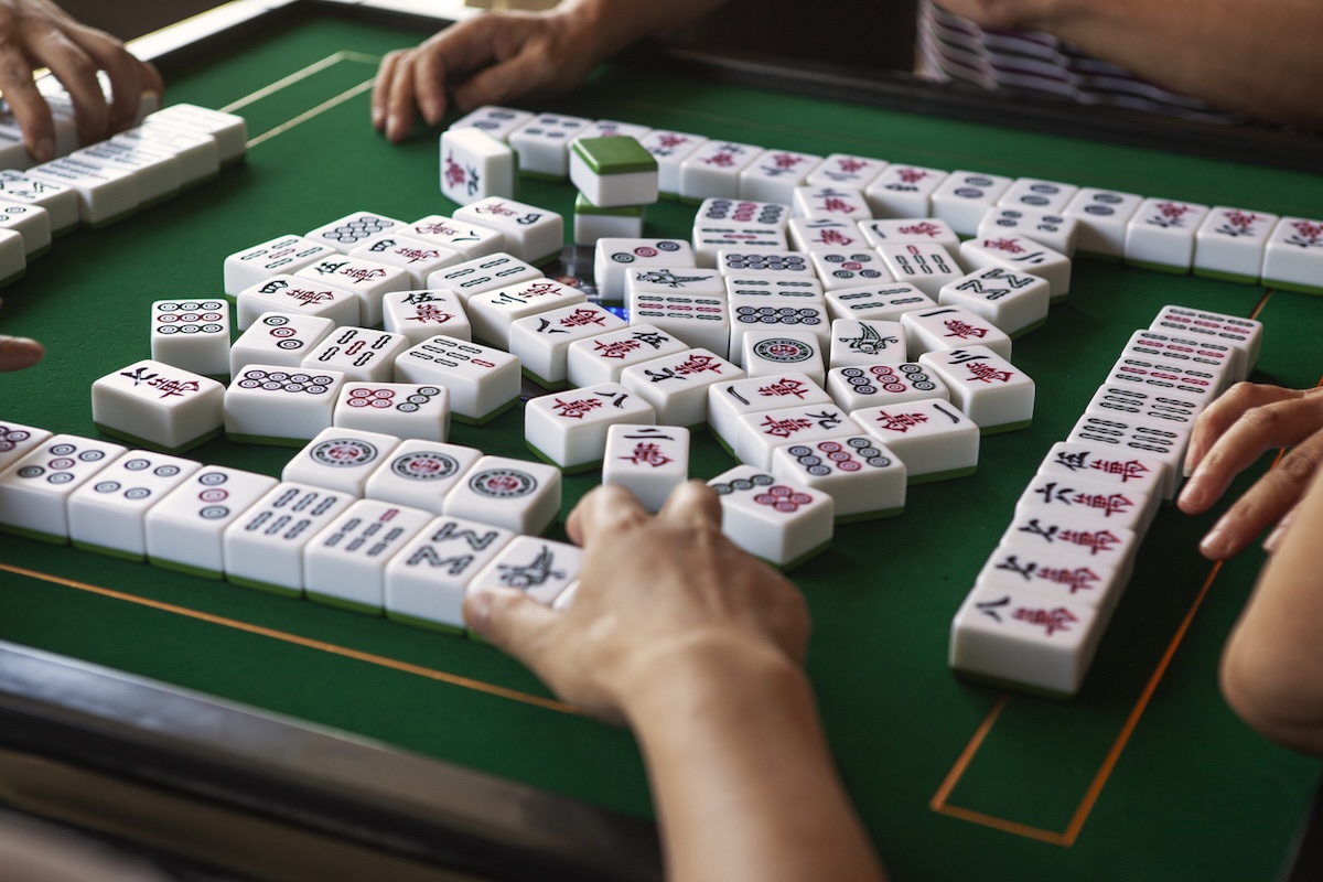 Four people enjoy a game of Mahjong on a green table, hands reaching for tiles in a scene that captures the relaxation of Mahjong retreats often found in the South, with rows of tiles arranged before each player.