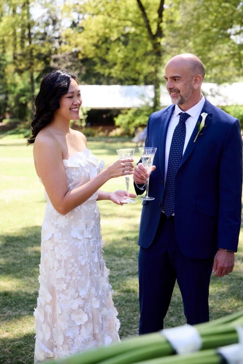 Peek Inside FIVE Dreamy Dixon Gallery & Gardens Weddings - 5 A woman in a white floral dress and a man in a blue suit clink glasses at Dixon Gardens on a sunny day, with love in the air.
