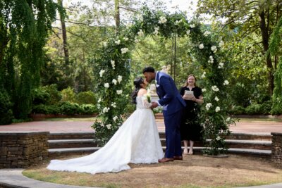 A bride and groom share a kiss at an outdoor wedding ceremony under a floral arch at Dixon Gardens, with an officiant standing behind them.