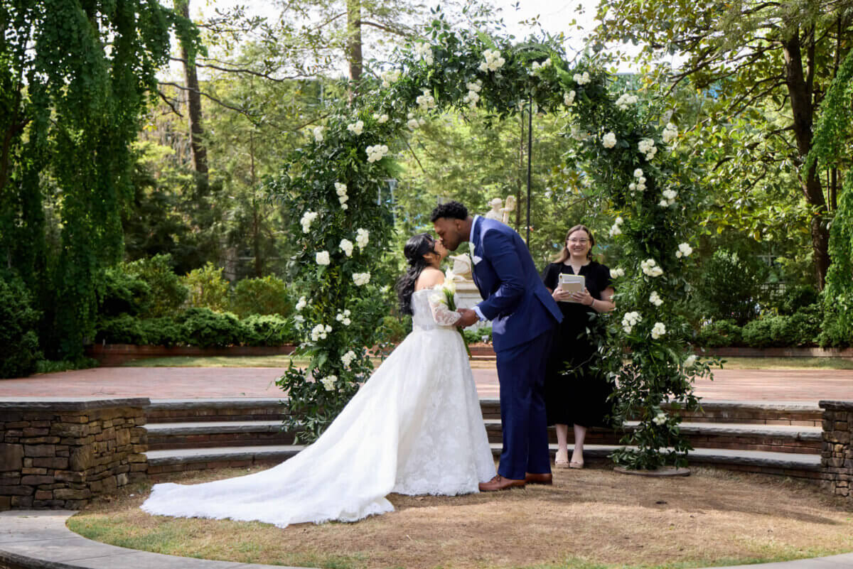 A bride and groom share a kiss at an outdoor wedding ceremony under a floral arch at Dixon Gardens, with an officiant standing behind them.