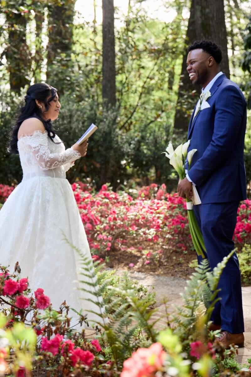 Peek Inside FIVE Dreamy Dixon Gallery & Gardens Weddings - 3 A bride in a white dress reads from a book to a groom in a blue suit holding calla lilies, standing in a garden with pink flowers and tall trees.