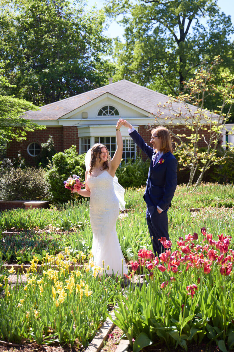 Peek Inside FIVE Dreamy Dixon Gallery & Gardens Weddings - 6 A bride and groom stand in the lush Dixon Gardens, with the groom twirling the bride by the hand. The bride holds a bouquet, and a brick building from Dixon Gallery graces the background.