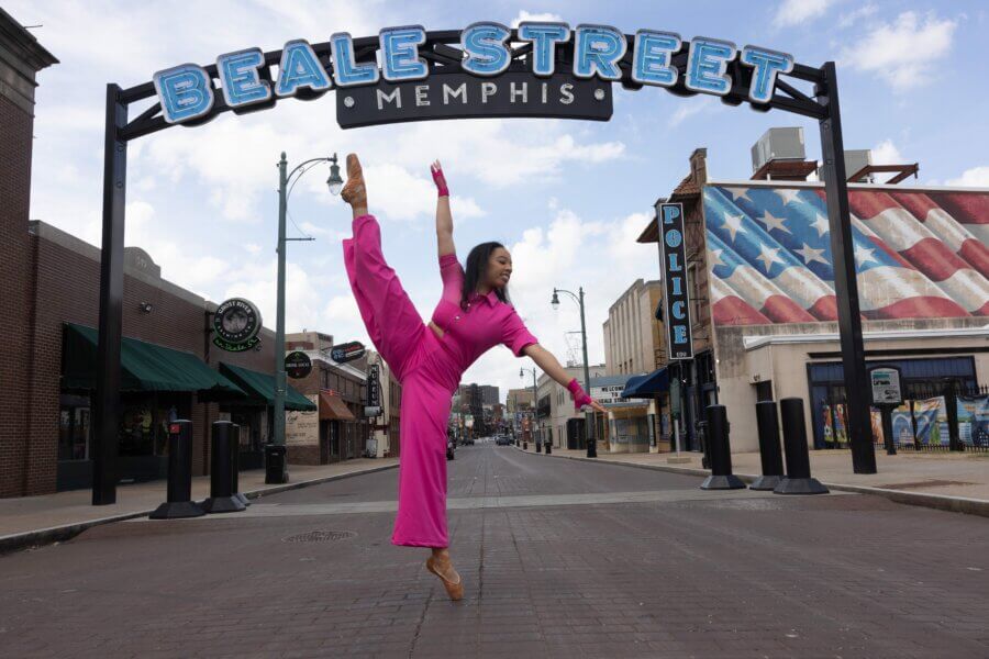Ashley Hannah Davis Brings Memphis to Life Through Dance - 6 Ashley Hannah Davis, dressed in a bright pink outfit, performs a ballet leap on Beale Street in Memphis, with an American flag mural in the background.