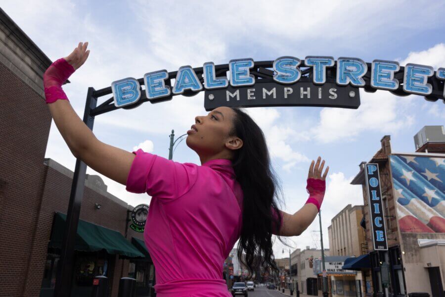 Ashley Hannah Davis Brings Memphis to Life Through Dance - 5 Ashley Hannah Davis, in a bright pink outfit, poses beneath the iconic Beale Street Memphis sign on a city street, with shops and a police station visible in the background.