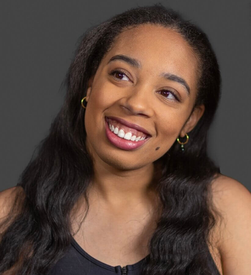 Ashley Hannah Davis Brings Memphis to Life Through Dance - 2 A woman with long dark hair and gold hoop earrings smiles, looking slightly to the side, against a plain dark background.