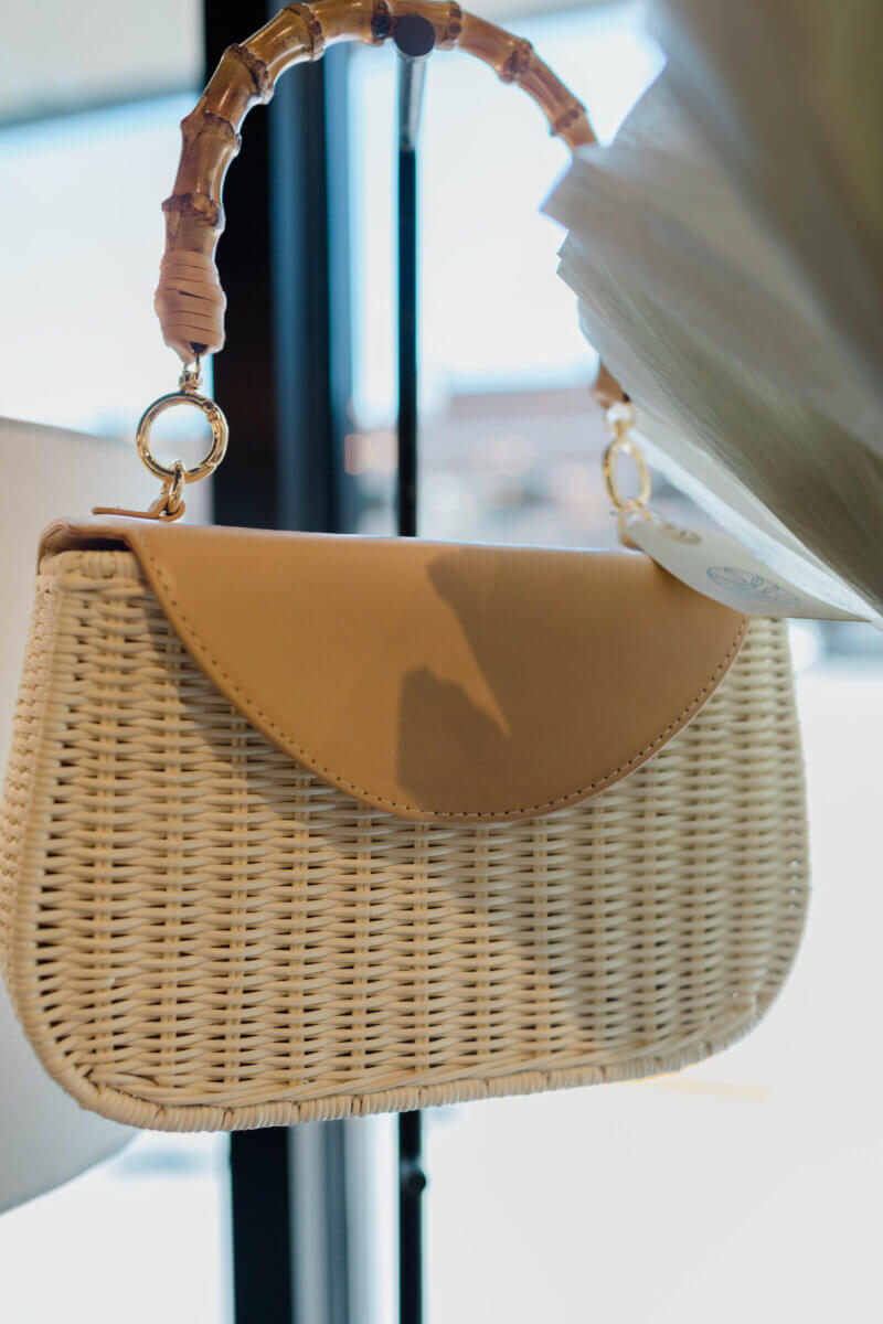 15 Memphis FINDS for May: Mother's Day & Spring Celebrations - 9 A woven handbag with a light tan flap and a curved bamboo handle hangs indoors near a window with sunlight shining through, perfect for your next Memphis shopping adventure.