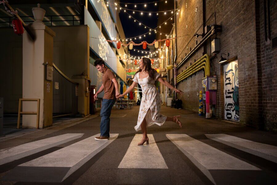 Why Hattiesburg Might Be Mississippi’s Biggest Surprise - 2 A man and woman cross a zebra-striped street in an alley at night, decorated with string lights and lanterns; the woman smiles and spins, while the man looks down.