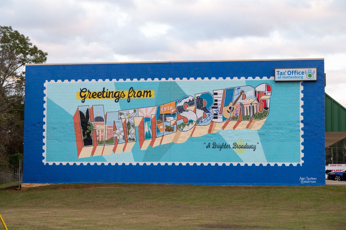 A colorful mural on a building in Hattiesburg, Mississippi, reads "Greetings from Hattiesburg" with images inside the letters and "A Brighter Gateway!" in cursive; Tax Office sign is visible above. This might be the city's biggest surprise!.