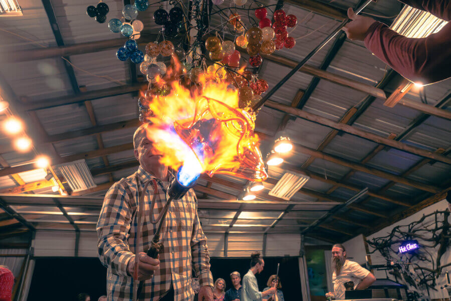Why Hattiesburg Might Be Mississippi’s Biggest Surprise - 9 A person in a plaid shirt shapes molten glass with a torch, creating a burst of flames, while onlookers in a Hattiesburg workshop watch the process beneath colorful glass art hanging above.