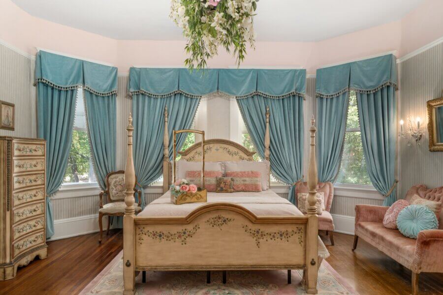 Why Hattiesburg Might Be Mississippi’s Biggest Surprise - 3 A vintage-style bedroom with a floral-painted wooden bed, blue curtains, floral arrangements, and pastel furniture, featuring ornate decor and hardwood floors.