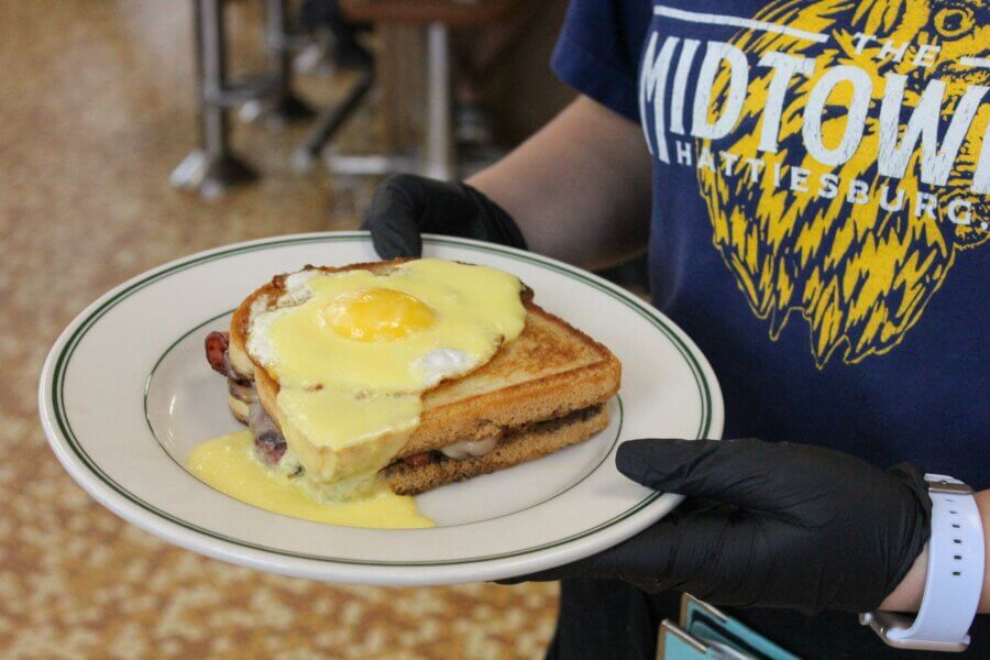 Why Hattiesburg Might Be Mississippi’s Biggest Surprise - 10 A person wearing black gloves holds a plate with a grilled sandwich topped with a fried egg and creamy yellow sauce, ready to enjoy a taste of Hattiesburg, Mississippi.