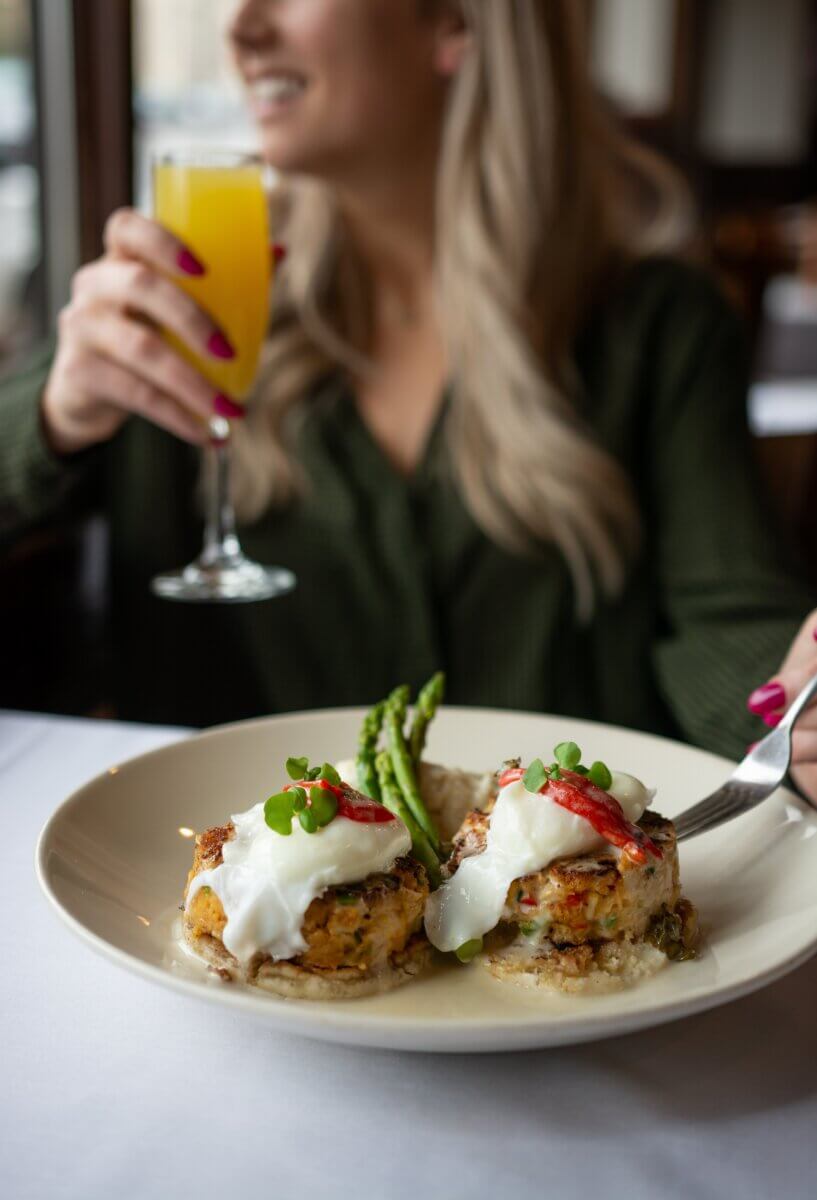 Where to Get Mother's Day Brunch in Memphis - 3 A woman holding a mimosa sits at a table during Mother's Day Brunch in Memphis, enjoying crab cakes topped with poached eggs, asparagus, and garnish.