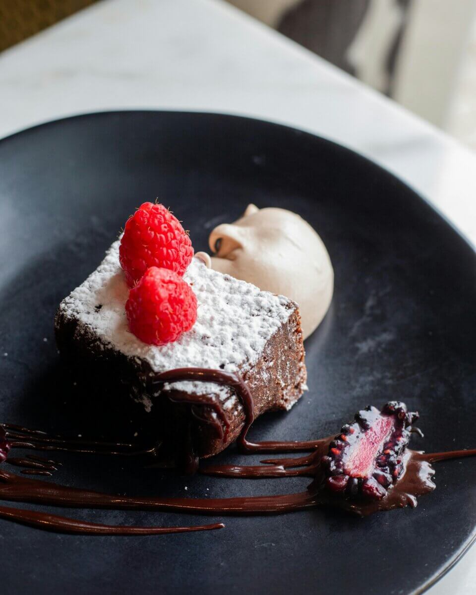 Where to Get Mother's Day Brunch in Memphis - 5 A square piece of chocolate cake topped with powdered sugar and raspberries, served with a dollop of cream and chocolate sauce on a black plate—perfect for a sweet treat at Mother's Day Brunch in Memphis.