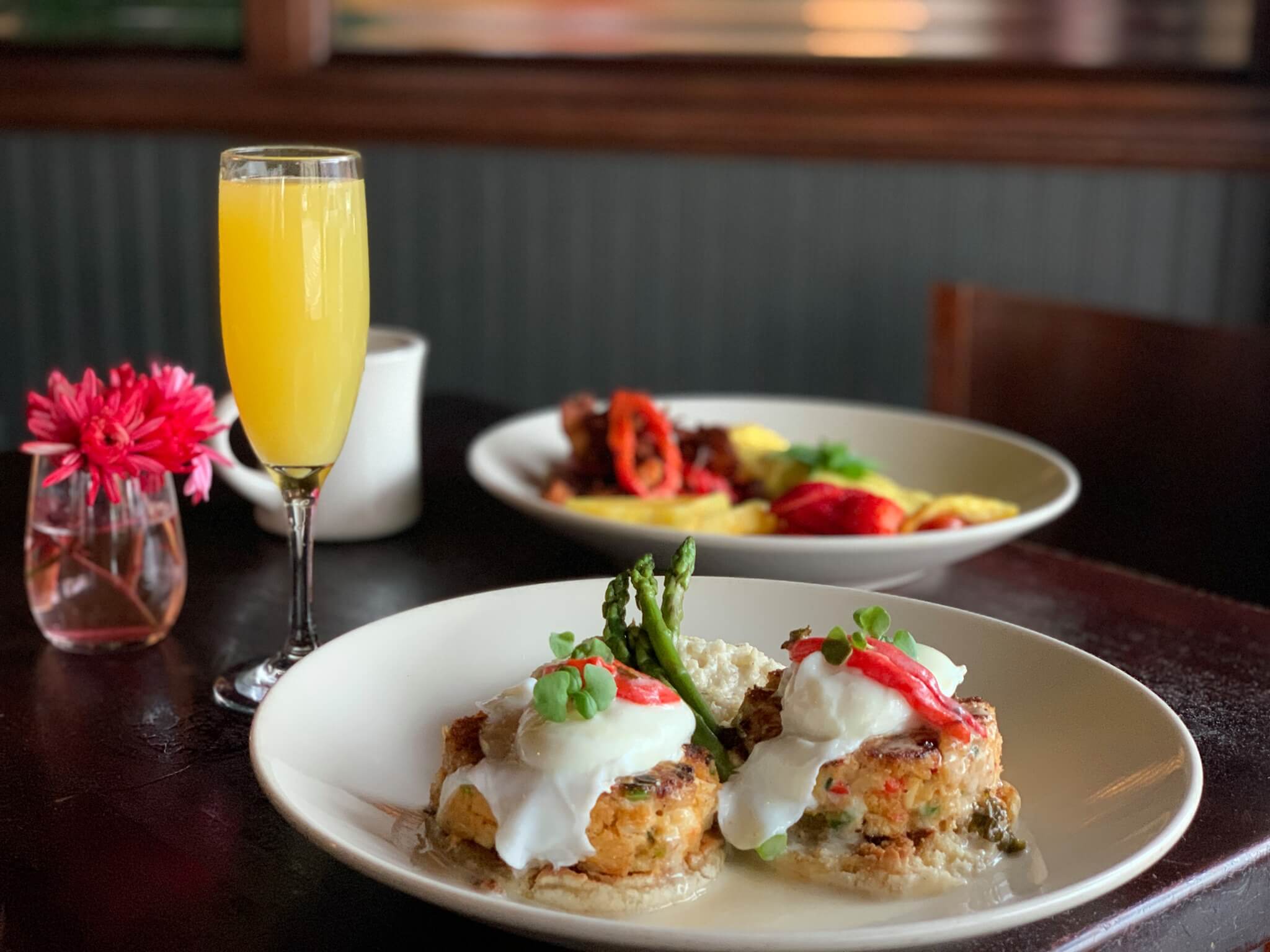 A plate with two crab cakes topped with poached eggs and asparagus sits next to a glass of orange juice—perfect for a Mother's Day Brunch in Memphis; another dish is blurred in the background.