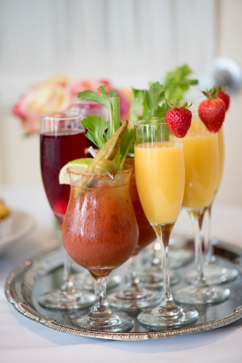 Where to Get Mother's Day Brunch in Memphis - 11 A silver tray holds various cocktails, perfect for Mother's Day Brunch in Memphis—a Bloody Mary with celery and okra, mimosas garnished with strawberries, and a red drink in champagne flutes. Flowers bloom in the background.