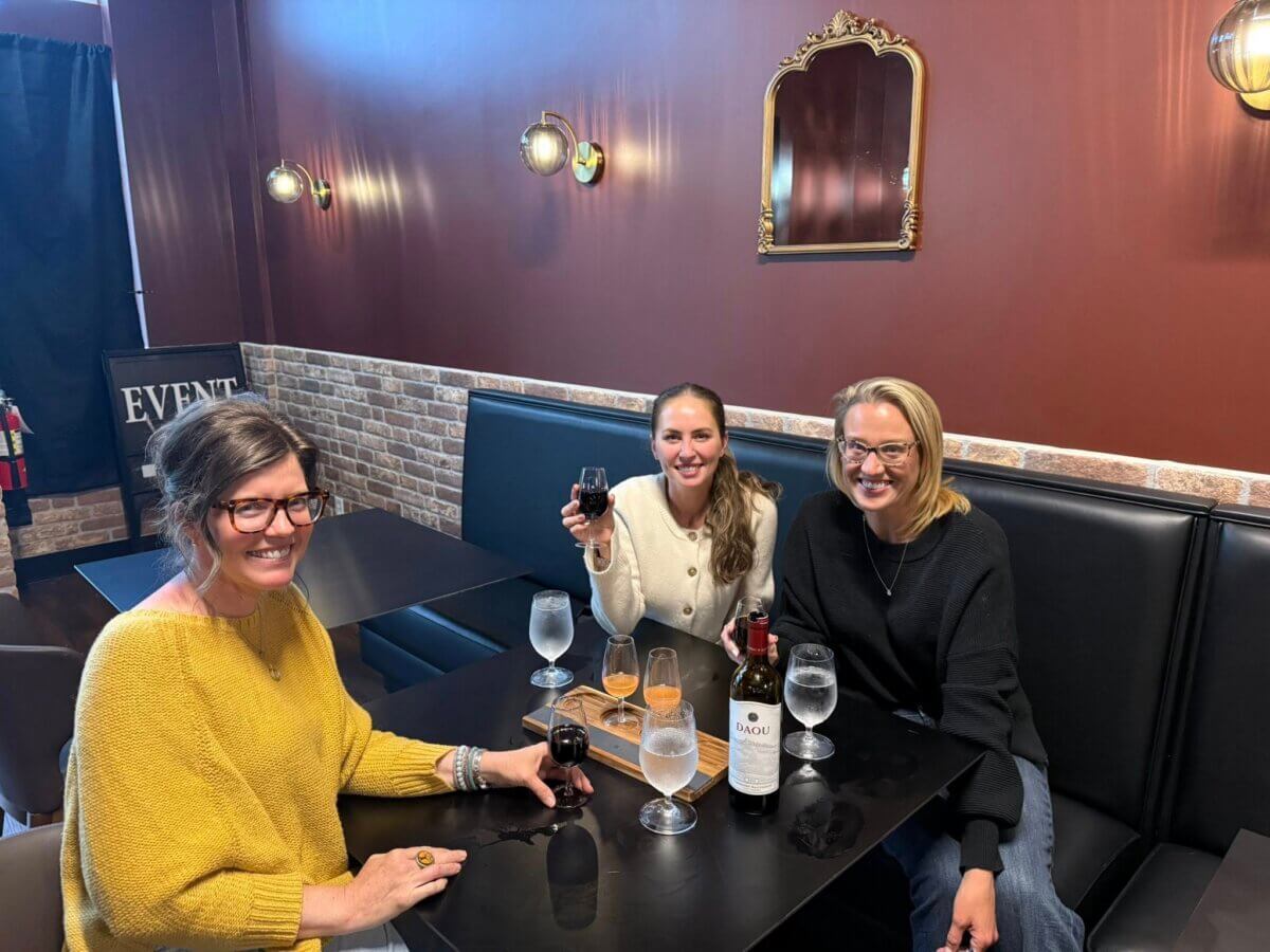 Three women sit at a restaurant table with wine, water glasses, and snacks, smiling at the camera. A mirror and wall lights are visible in the background, capturing the inviting vibe of new Memphis restaurants.