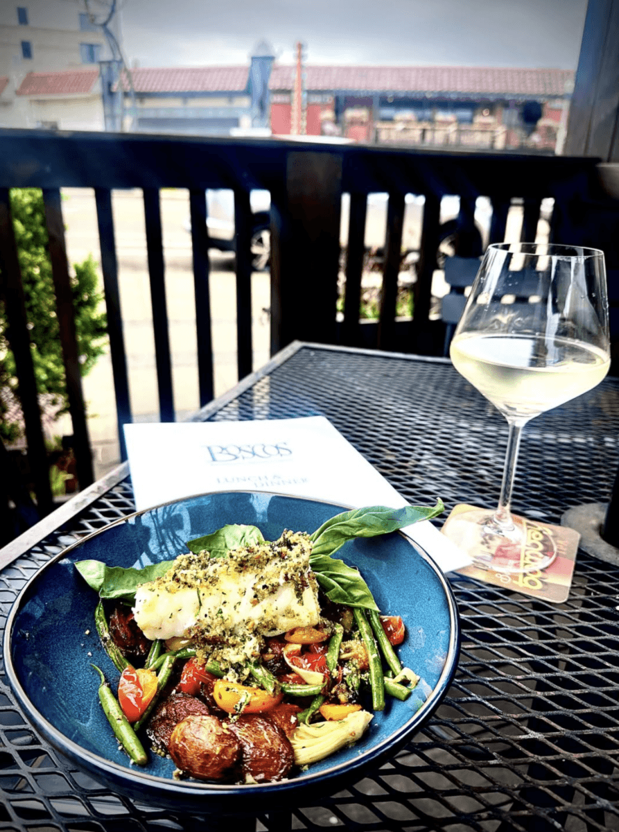 7 Memphis Patios Hosting Live Music This Season - 7 A plate of fish with roasted vegetables and greens next to a glass of white wine on an outdoor restaurant table; menu and street view in the background.