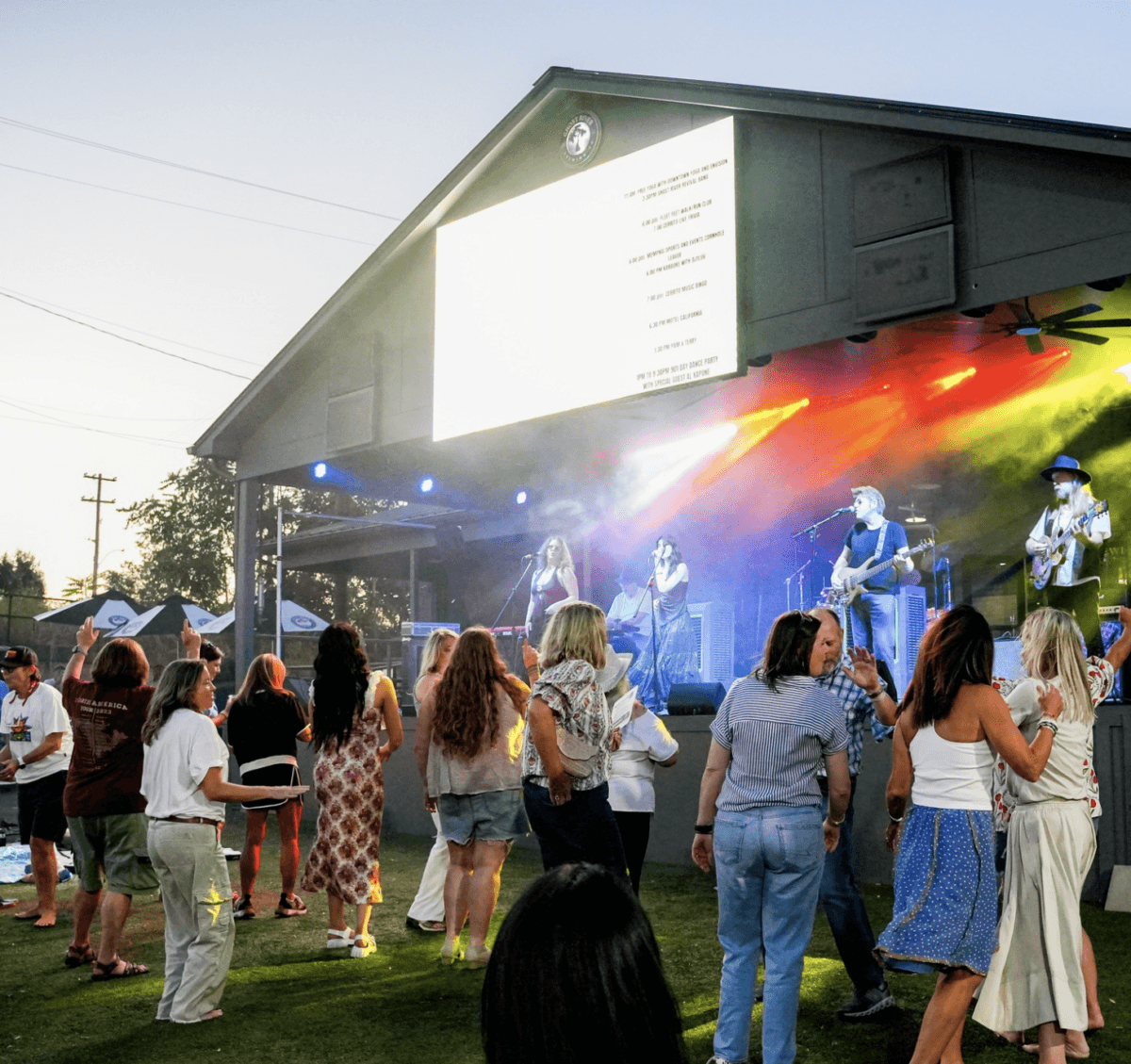 7 Memphis Patios Hosting Live Music This Season - 3 People dance on grass in front of an outdoor stage with a live band performing and colorful lights, during an evening event.