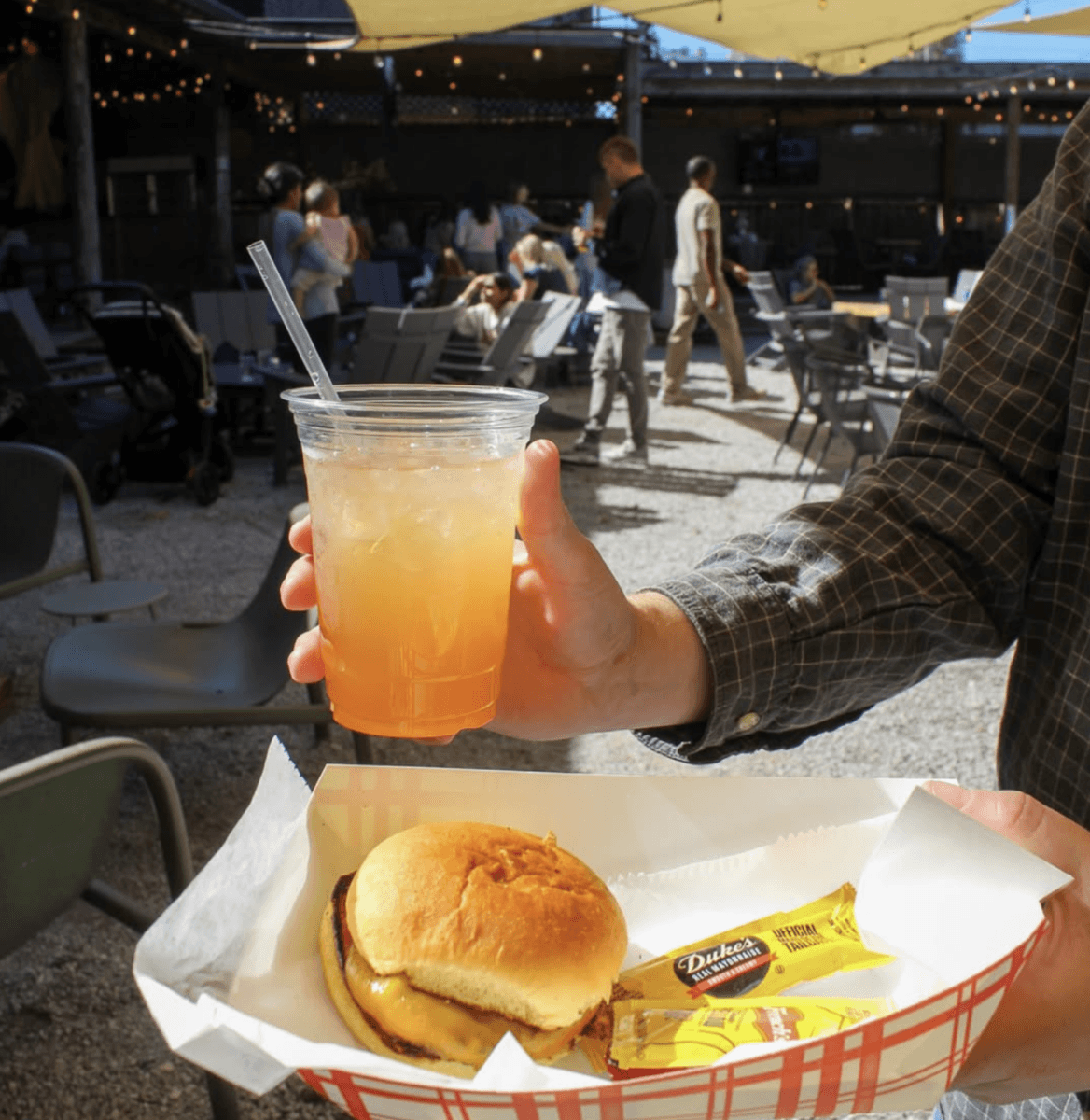 7 Memphis Patios Hosting Live Music This Season - 4 Person holding a tray with a cheeseburger and mustard packet in one hand and an iced drink in the other at an outdoor seating area with people in the background.