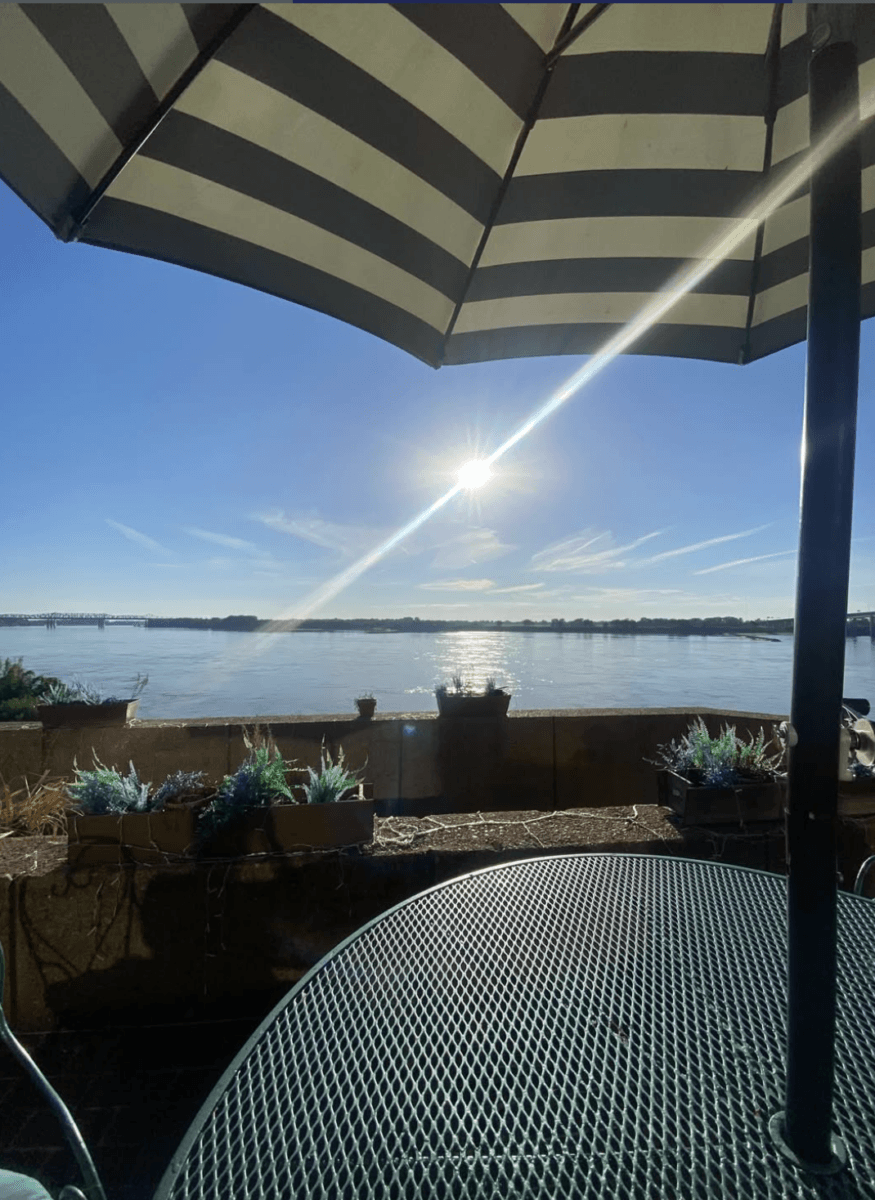 7 Memphis Patios Hosting Live Music This Season - 5 A striped umbrella shades a metal patio table overlooking a calm body of water under a bright sun and clear blue sky.