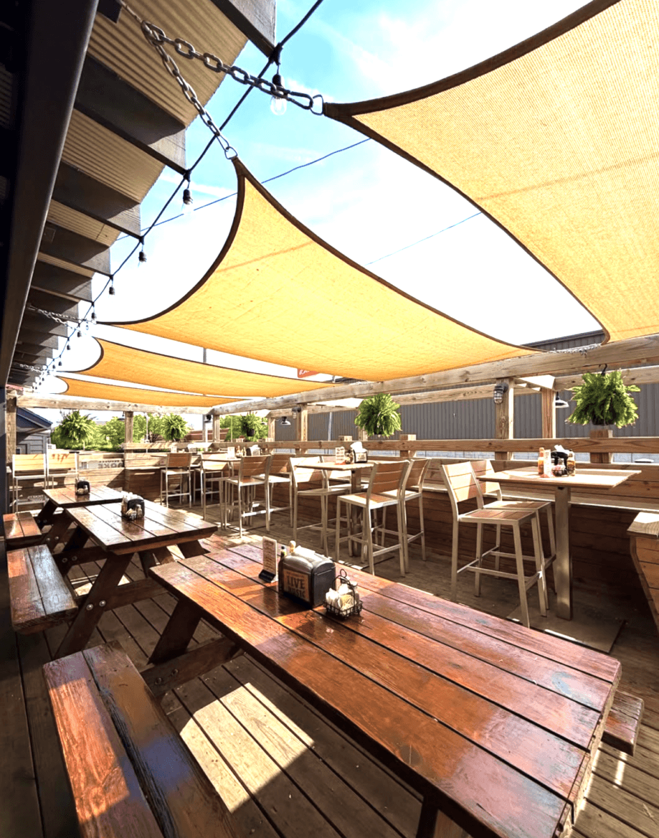 7 Memphis Patios Hosting Live Music This Season - 8 Outdoor patio with wooden picnic tables, high-top tables, hanging plants, and sunshades overhead on a sunny day.