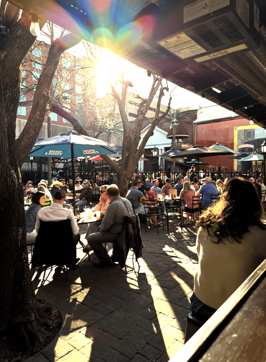 7 Memphis Patios Hosting Live Music This Season - 6 People sit at outdoor tables in a busy patio restaurant under umbrellas, with sunlight streaming through trees and casting long shadows across the scene.