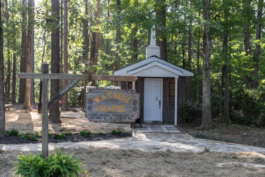 Hidden History in the Southβs Most Beautiful State Parks - 10 Small brick chapel with a white door and steeple surrounded by trees in one of the most beautiful state parks. A wooden sign in front reads "Wayside Chapel" with an inspirational quote below.