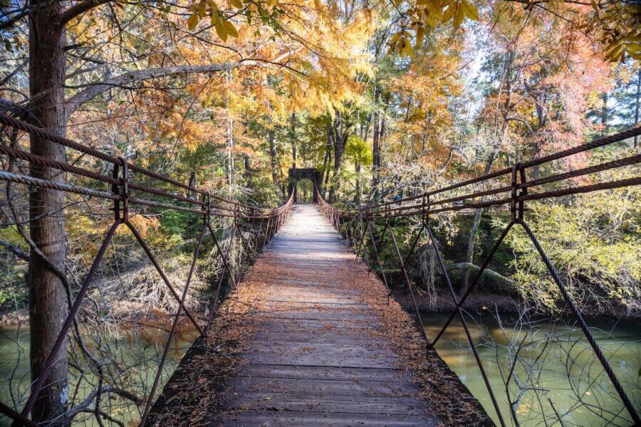 Hidden History in the Southβs Most Beautiful State Parks - 8 A wooden suspension bridge with metal railings spans over a river, surrounded by autumn trees with orange and yellow leaves.