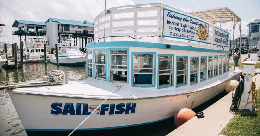 Your Guide to Coastal Mississippi’s Seafood Scene - 6 A white boat named "SAIL FISH" is docked at a marina, with a large sign advertising fishing charter services on its upper deck—a perfect starting point for exploring MS activities and sampling local Coastal MS cuisine. Other boats and buildings are in the background.