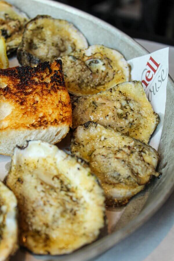 Your Guide to Coastal Mississippi’s Seafood Scene - 9 A metal plate with grilled oysters topped with herbs and a slice of toasted bread on parchment paper, capturing the essence of Coastal MS cuisine.