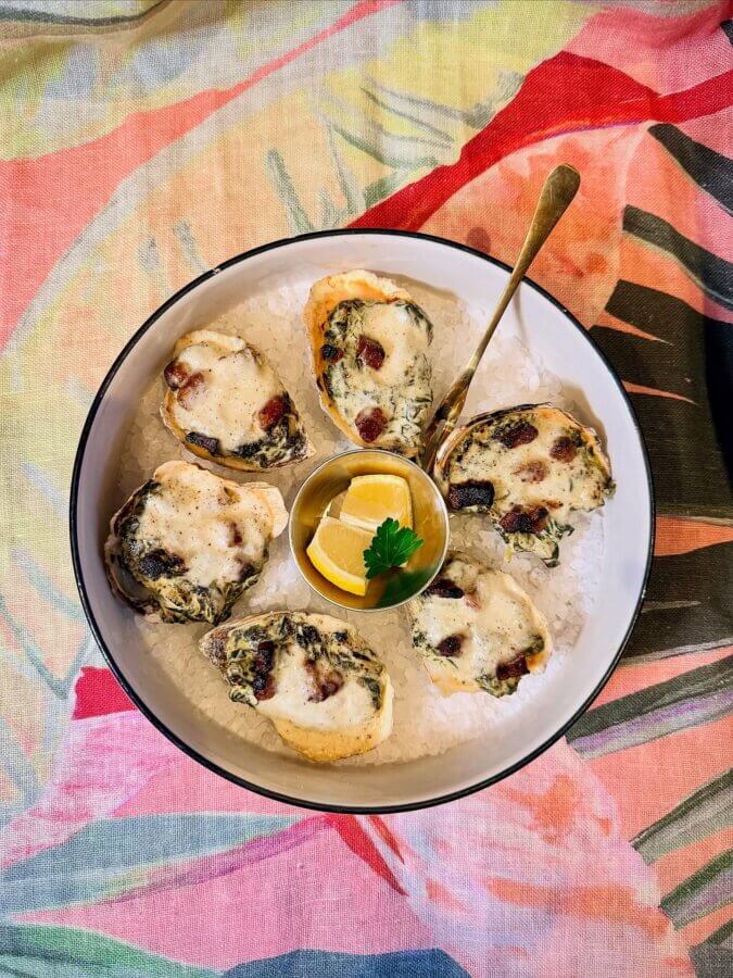 Your Guide to Coastal Mississippi’s Seafood Scene - 2 A bowl of baked oysters topped with a creamy sauce and herbs, served on ice with lemon wedges and a garnish, highlights the Coastal MS Cuisine on a colorful patterned tablecloth.