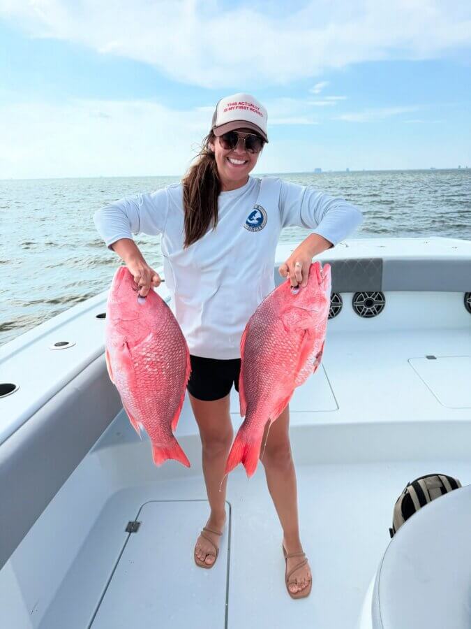 Your Guide to Coastal Mississippi’s Seafood Scene - 4 A woman stands on a boat holding two large red fish, smiling at the camera, enjoying one of the top activities along the ocean—perfect for lovers of Coastal MS cuisine and fresh catches.