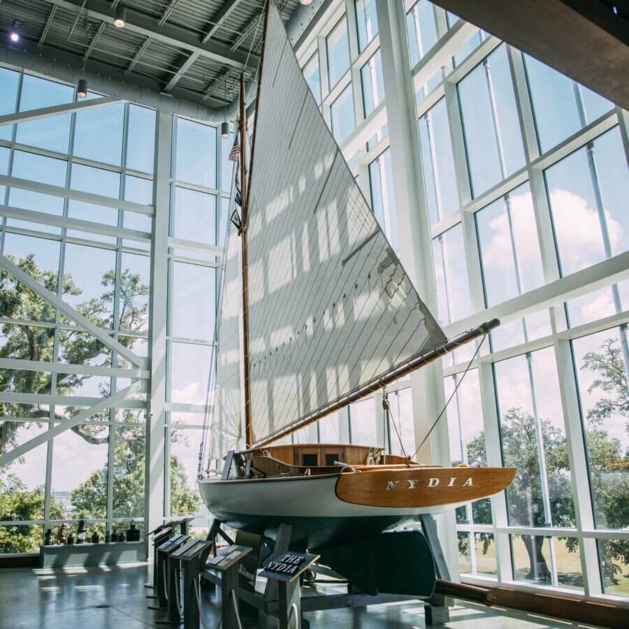 Your Guide to Coastal Mississippi’s Seafood Scene - 5 A wooden sailboat named NYDIA is displayed indoors on stands, surrounded by large glass windows with trees and blue sky visible outside—a scene capturing the relaxed charm of the Mississippi coast and its inviting seafood spots.