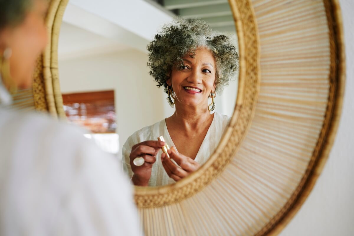 The Facelift Has Gotten a Major Rebrand - 3 An older woman with curly gray hair smiles at her reflection in a round mirror while holding a lipstick tube, embracing her beauty after a recent facelift.