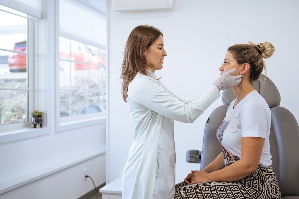 The Facelift Has Gotten a Major Rebrand - 2 A healthcare professional wearing gloves examines the face of a seated woman in a medical office, assessing her suitability for a facelift or other cosmetic surgery.