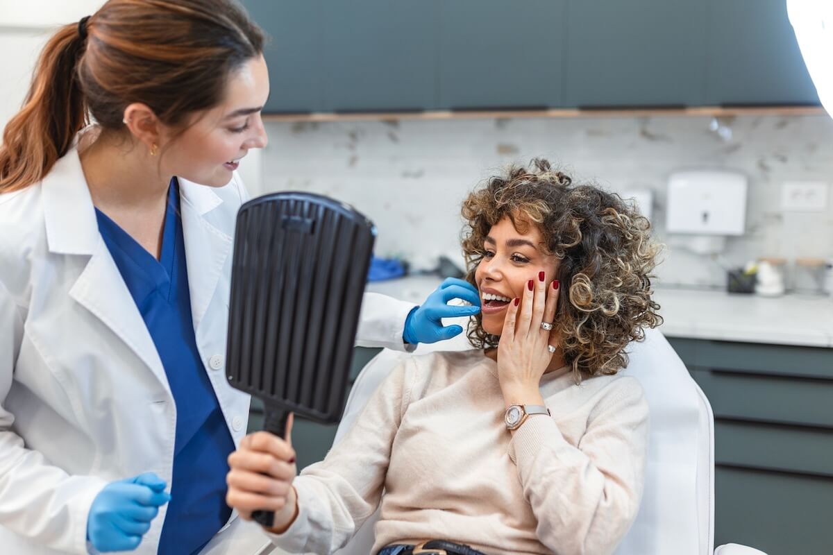 The Facelift Has Gotten a Major Rebrand - 4 A woman looks at her reflection in a hand mirror, while a technician or nurse in a white coat and blue gloves discusses facelift options and with her.