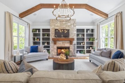 Bright living room at 2709 Edlington Terrace features a stone fireplace, wooden mantel, wall-mounted TV, built-in shelves, beige sofas with blue accents, a round coffee table, and exposed ceiling beams.