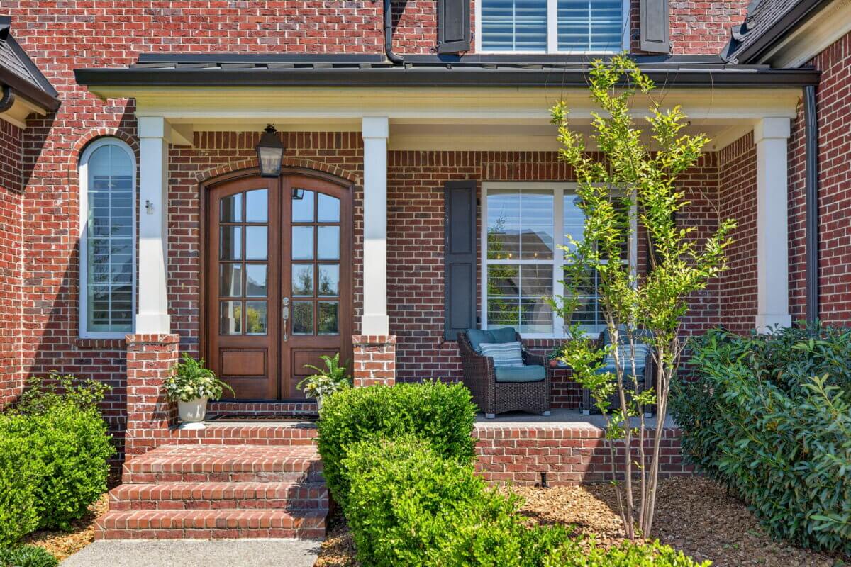 Just Listed! A Thoughtfully Designed Franklin Home That Delivers on Every Level - 3 Charming brick house at 2708 Eglington Terrace features a front porch with two columns, arched wooden door, windows with shutters, potted plant, cushioned outdoor chair, and landscaped bushes.
