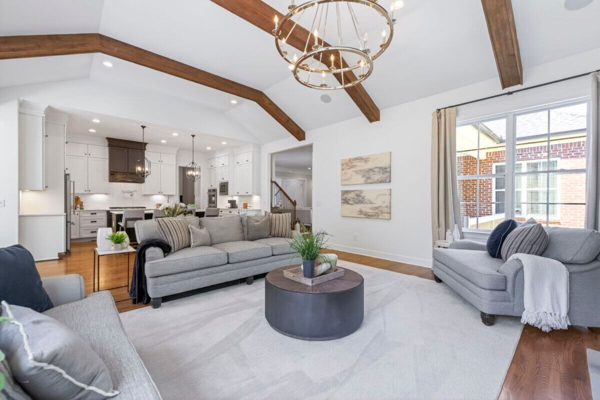 Just Listed! A Thoughtfully Designed Franklin Home That Delivers on Every Level - 7 Spacious living room features gray sofas, a round coffee table, exposed wooden ceiling beams, and an open view into a modern white kitchen.