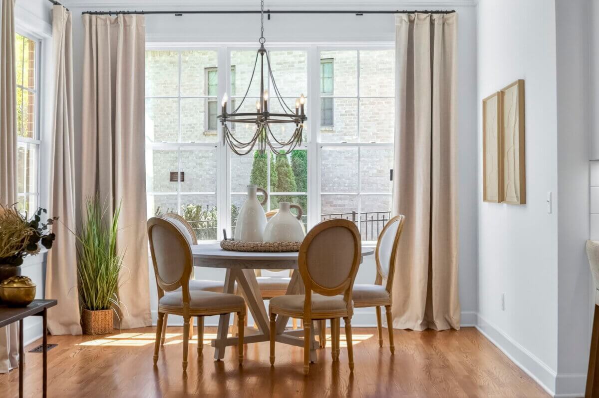 Just Listed! A Thoughtfully Designed Franklin Home That Delivers on Every Level - 9 A round dining table with four upholstered chairs sits by large windows with beige curtains; a chandelier hangs above and decorative vases are on the table.
