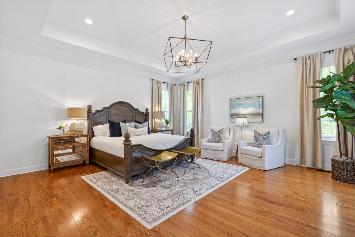 Just Listed! A Thoughtfully Designed Franklin Home That Delivers on Every Level - 10 Spacious bedroom at 2708 Eglington Terrace featuring a large bed, two white armchairs, a patterned rug, wood floors, and a modern chandelier. Natural light pours through windows adorned with beige curtains.
